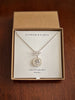 A silver necklace with a toggle clasp and a coin charm pendant presented in a brown kraft gift box on a cream card labeled 'FLOWER & FABLE' and 'FORGET ME NOT'.