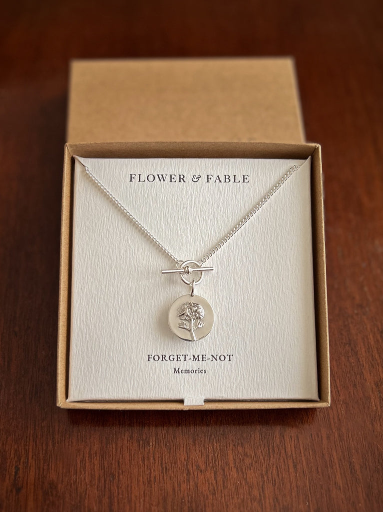 A silver necklace with a toggle clasp and a coin charm pendant presented in a brown kraft gift box on a cream card labeled 'FLOWER & FABLE' and 'FORGET ME NOT'.