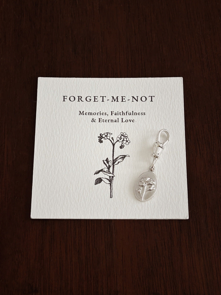 Sterling Silver Forget-Me-Not flower medal with a secure clasp on a matching card with text on a wooden surface.