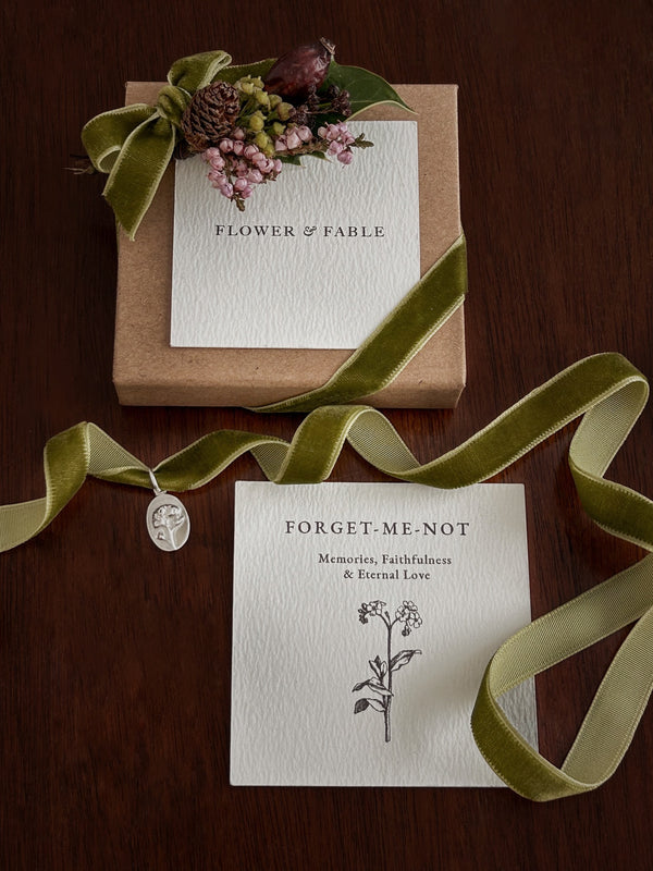 Floral arrangement with 'Flower & Fable' branding on a wooden surface and a forget me not charm