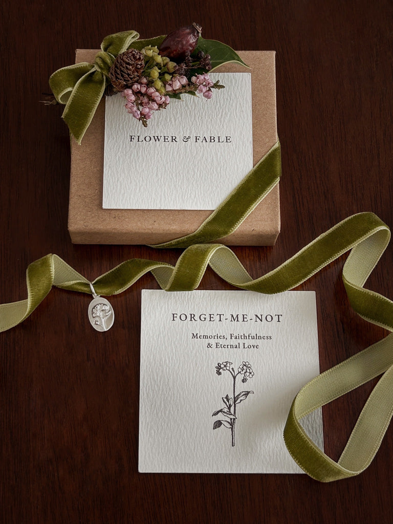 Floral arrangement with 'Flower & Fable' branding on a wooden surface and a forget me not charm