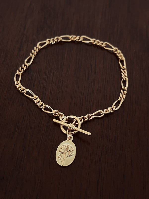 Gold forget me not charm bracelet with figaro chain and t bar closure on a dark wooden surface