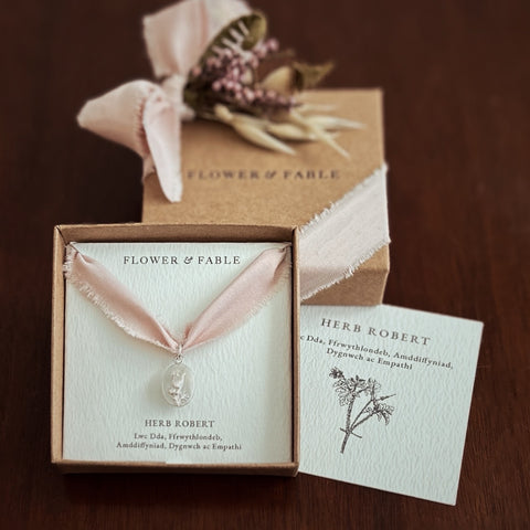 Herb Robert Charm Necklace with pink ribbon and 'Flower & Fable' packaging on a wooden surface