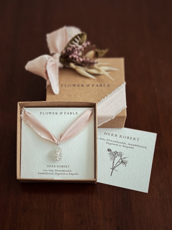 Herb Robert Charm Necklace with pink ribbon and 'Flower & Fable' packaging on a wooden surface