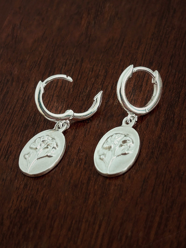 Pair of sterling silver huggie hoop earrings with forget me not medal charms on a dark wooden surface