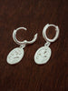 Pair of sterling silver huggie hoop earrings with forget me not medal charms on a dark wooden surface