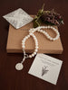 Pearl necklace with a silver lily pendant on a brown box with a card and flowers.