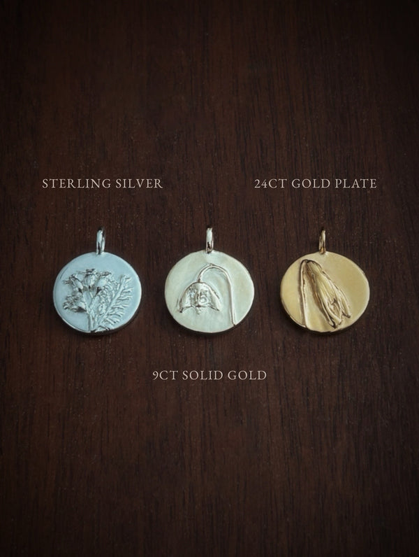 Three gold pendants on a wooden surface labeled 'Sterling Silver', '24ct Gold Plate', and '9ct Solid Gold'.
